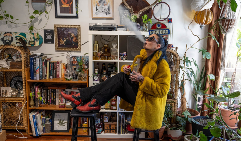 A woman sitting in a vintage room exhaling from a glass bong