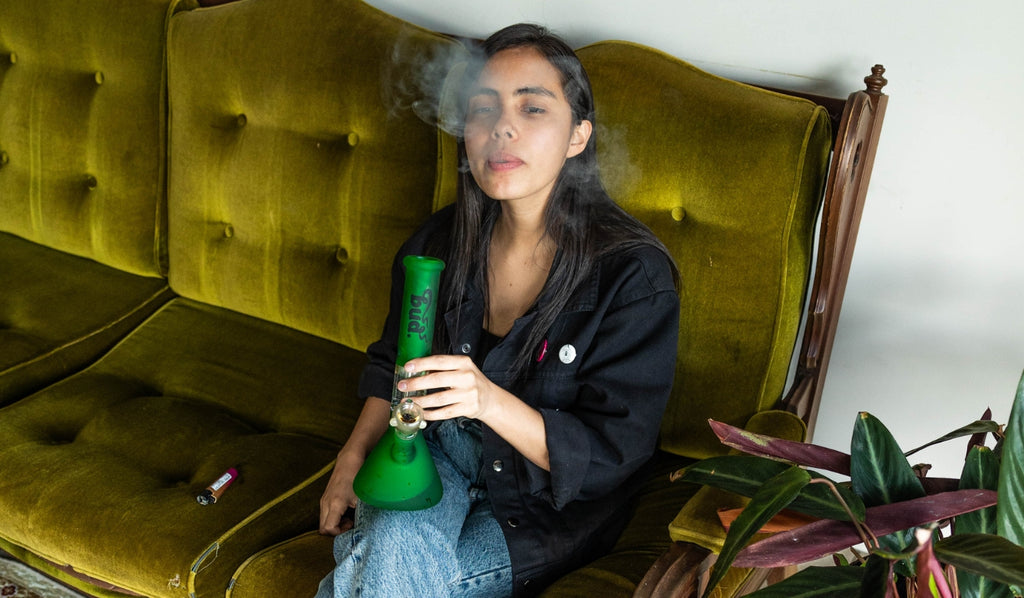 A woman sitting on a couch exhaling from a beaker glass bong