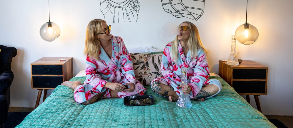 Two women sitting on a bed with a Her Highness Bong